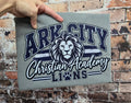 Heather gray Comfort Colors short sleeve tee featuring Ark City Christian Academy Lions in navy and white