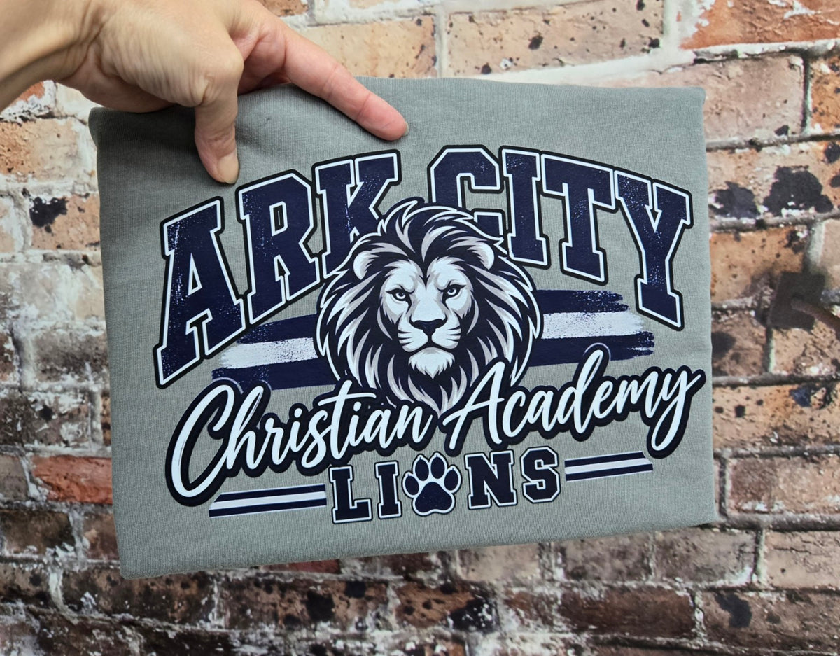 Heather gray Comfort Colors short sleeve tee featuring Ark City Christian Academy Lions in navy and white
